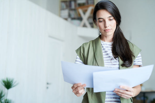 A woman in a bright home interior reads through paper documents, comparing pages as she reviews them