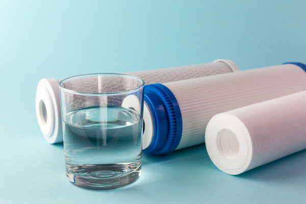 Glass of clear drinking water beside three water filter cartridges on a light blue surface