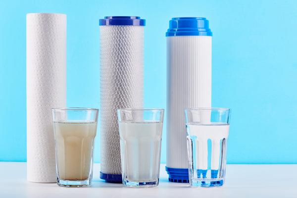 Three water filter cartridge types, a sediment depth filter, a carbon block, and a granular activated carbon cartridge, standing behind glasses of water filtered from dirty to clear