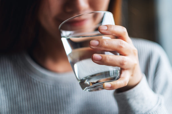 Woman in a grey sweater holding a clear glass of water to her mouth, about to take a sip