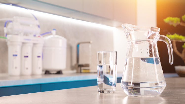 Glass and pitcher of filtered water on a kitchen counter with an under-sink reverse osmosis system and storage tank in the background
