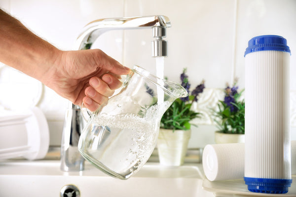 Clear glass pitcher being filled with water at a home kitchen faucet beside a water filter cartridge