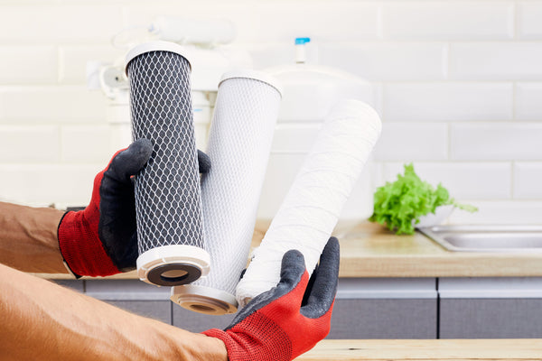 Gloved hands holding replacement sediment and carbon block water filter cartridges in a kitchen