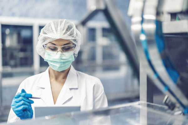 Technician in a hairnet, face mask and safety glasses reviewing a tablet beside stainless production equipment in a clean facility