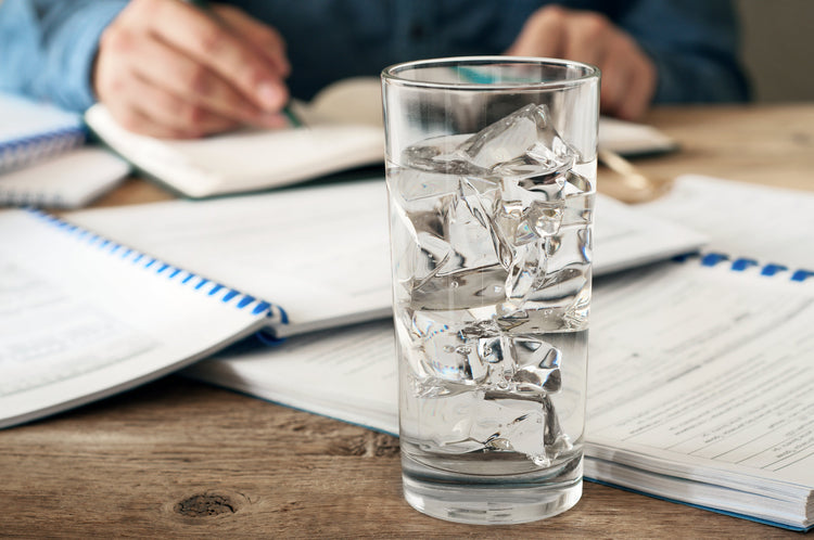 Tall glass of ice water on a wooden table beside open notebooks while a person writes in the background