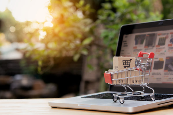 Laptop with a miniature shopping cart and box on a wooden surface, blurred greenery in the background
