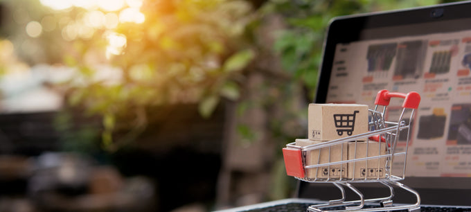 Laptop with a miniature shopping cart and box on a wooden surface, blurred greenery in the background