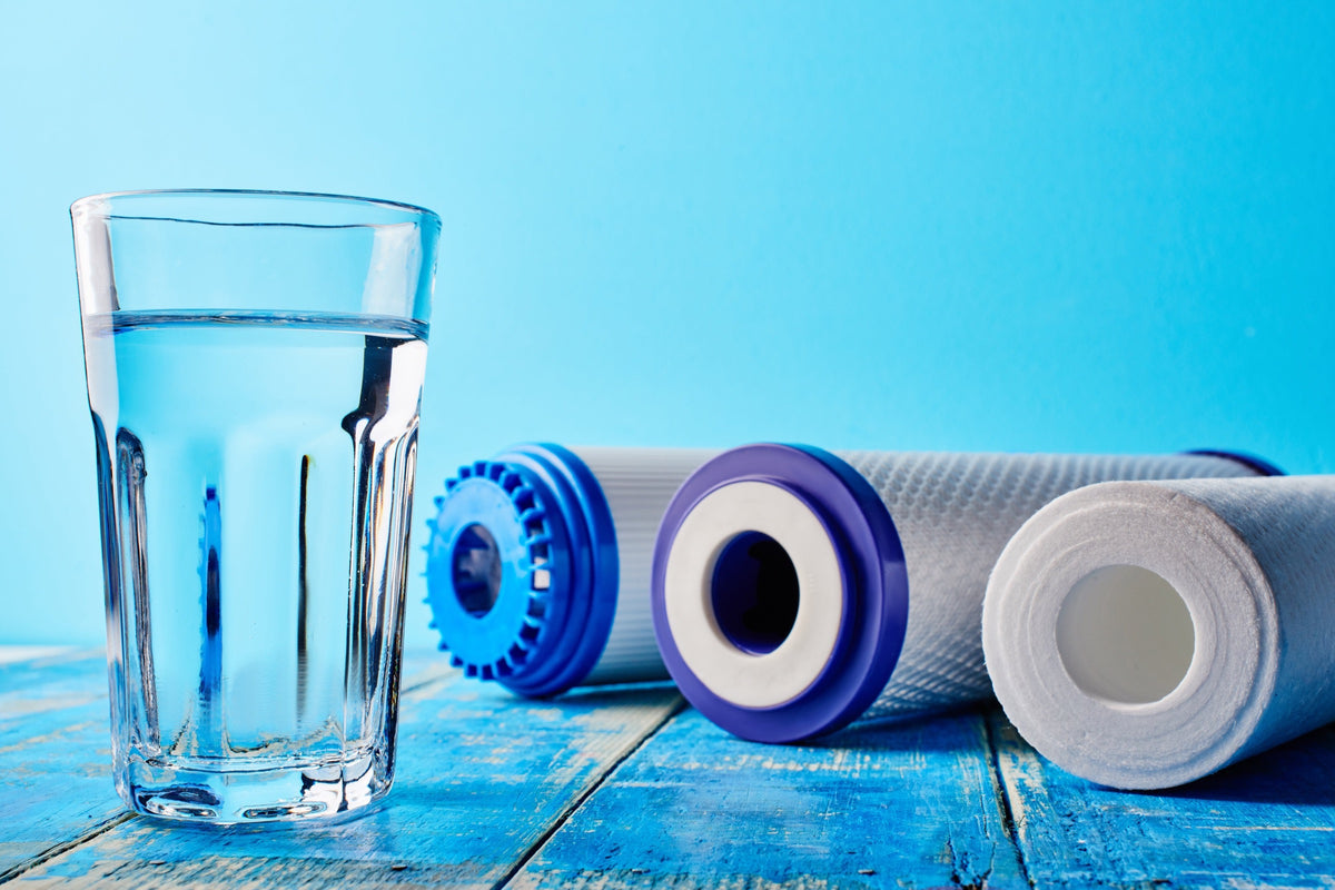Glass of clear filtered water beside three water filter cartridges on a blue wood table