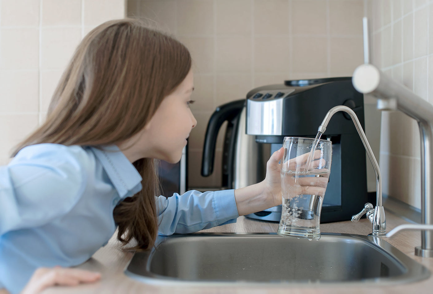 Child filling a glass of drinking water from a residential filtered faucet at the kitchen sink, illustrating what reverse osmosis removes from tap water