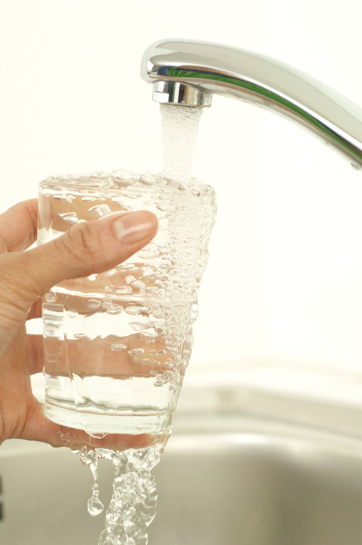 A hand fills a clear drinking glass with tap water from a chrome kitchen faucet over a stainless steel sink