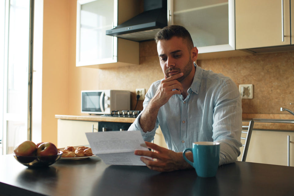 Man reading a printed letter at his kitchen table beside a teal mug, with a kitchen faucet in the background