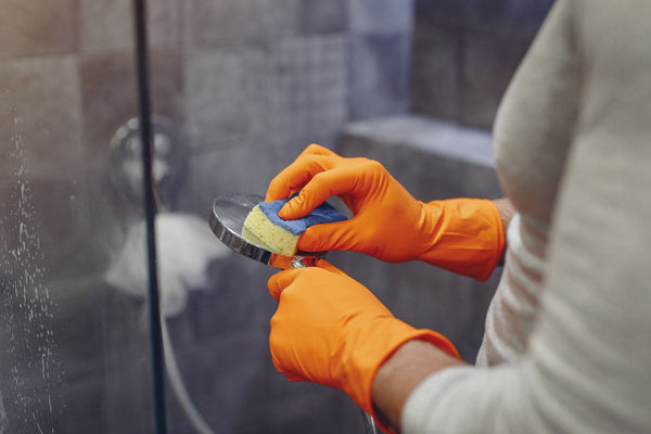 Gloved hands scrubbing a chrome shower head with a sponge in a home shower, with water spots on the glass door