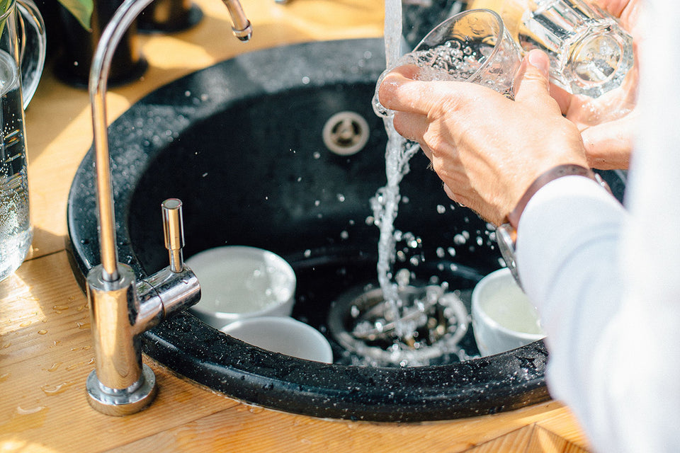 Water pouring into a dark round kitchen sink as someone rinses drinking glasses beside a slim gooseneck faucet