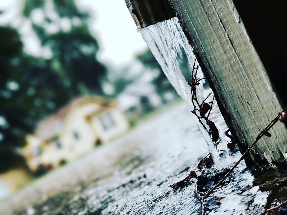 Rainwater pours off a roof edge carrying dead leaves and twigs, with a blurred house and trees behind