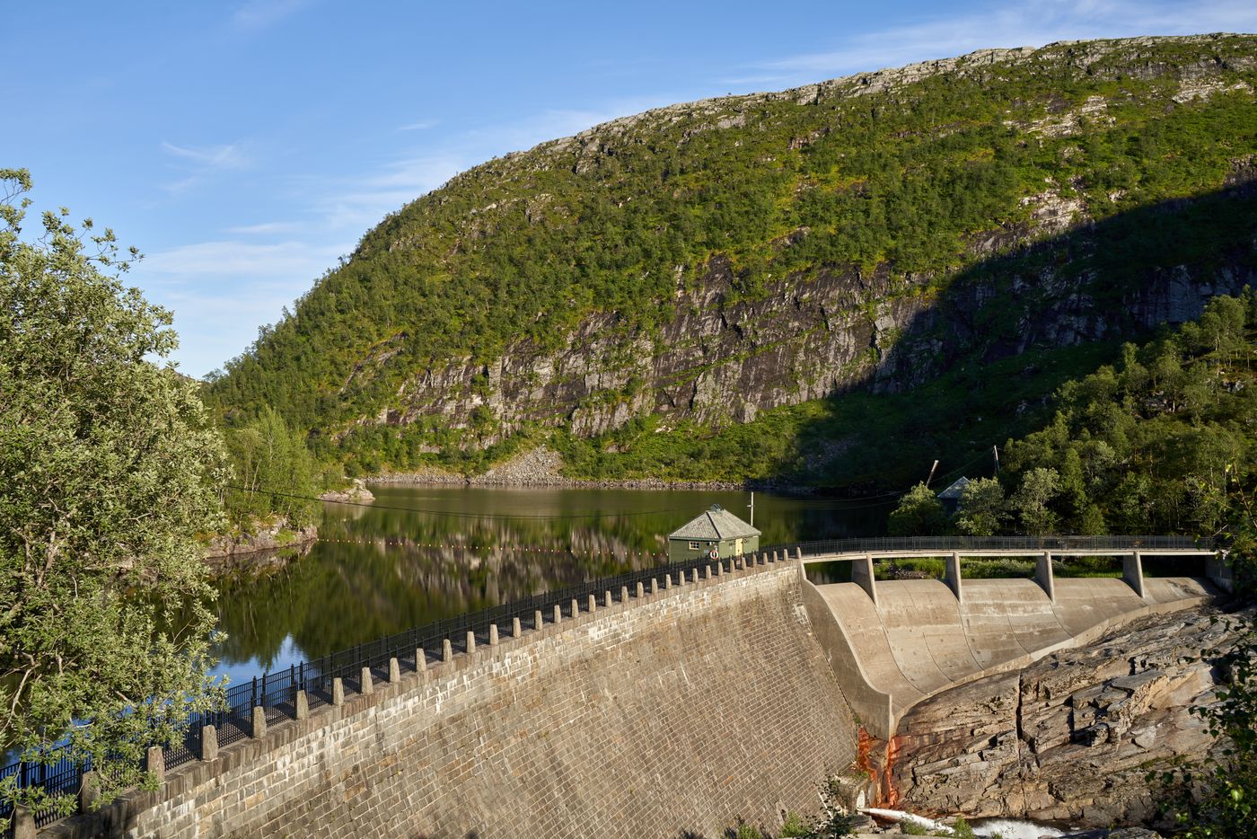 A stone reservoir dam holding back a calm lake below a forested mountain, the kind of surface-water source where harmful algal blooms form