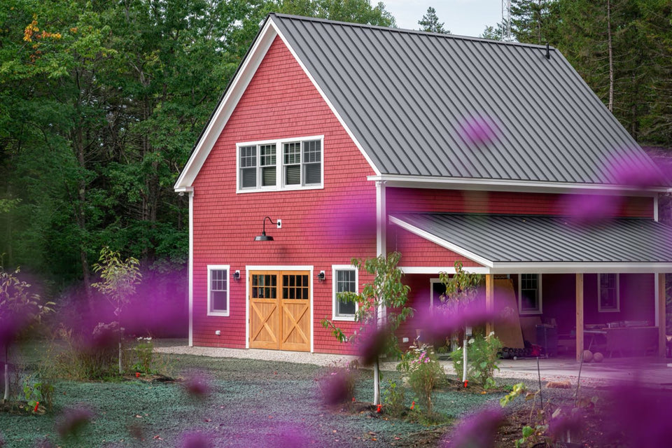 Red country home with a gray metal roof and wooden carriage doors on a wooded lot with a newly seeded yard