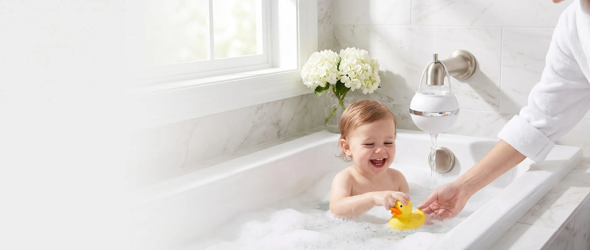 A parent and child playing in a bubble bath while a white Bath Ball hangs on the spout