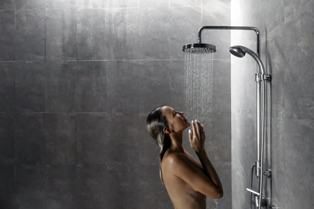 Woman tilting her head back under a rainfall shower head in a gray-tiled bathroom